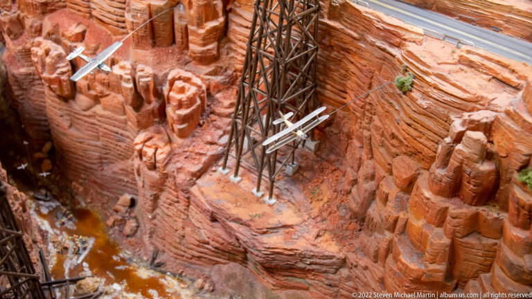 Dark trestle bridge spans a red-rock miniature canyon beside a small white aircraft.