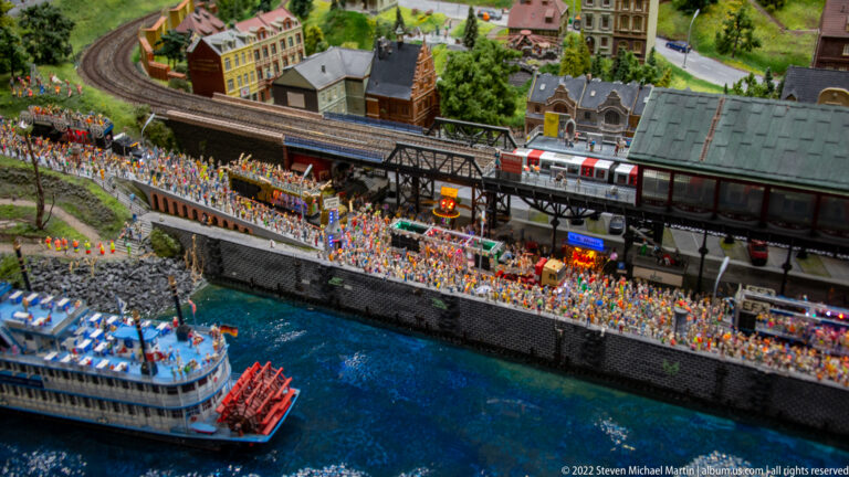 Excursion boat passes a crowded waterfront below an elevated station with a red-and-silver model train.