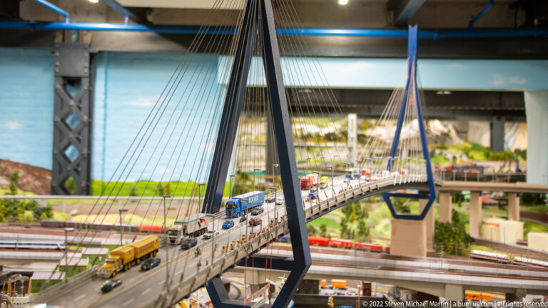 Miniature cable-stayed highway bridge with cars and trucks above red trains on tracks below.
