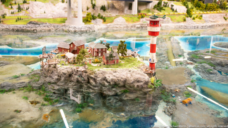 Red-and-white miniature lighthouse and small houses stand on a rocky islet surrounded by blue water.