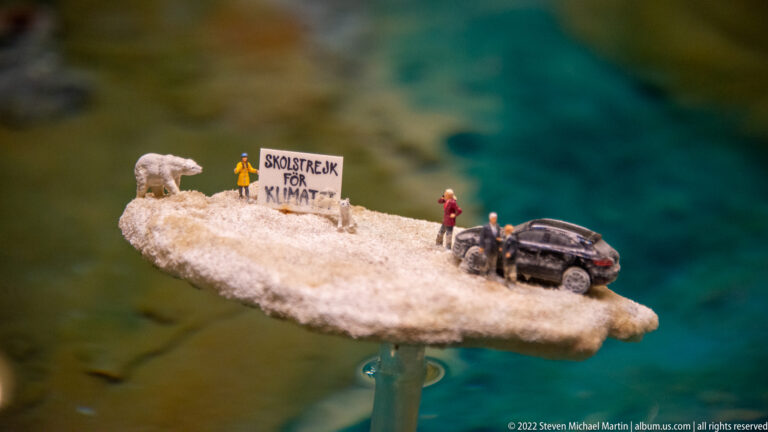 Miniature figures, a dark car, and a polar bear-like figure stand beside a “SKOLSTREJK FÖR KLIMAT” sign on a small ice floe.
