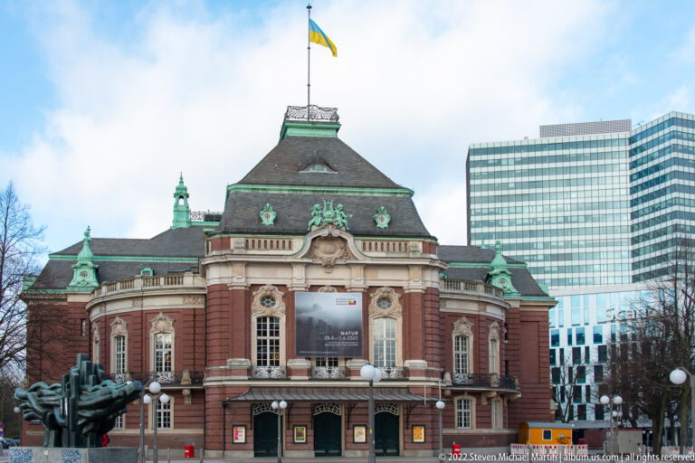 Red-brick Laeiszhalle in Hamburg with ornate stone entrance, green roof details, and modern office towers behind.