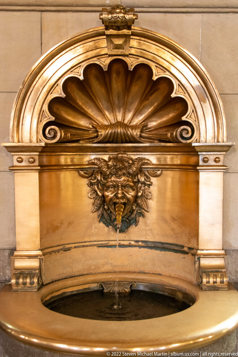 Polished brass wall fountain with scallop-shell hood, grotesque face spout, and shallow basin framed by pale stone.