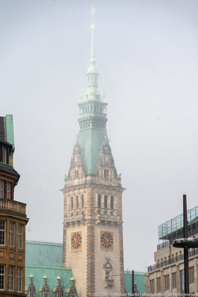 Hamburg Hauptbahnhof clock tower with twin clock faces and green copper roof rising through light haze.
