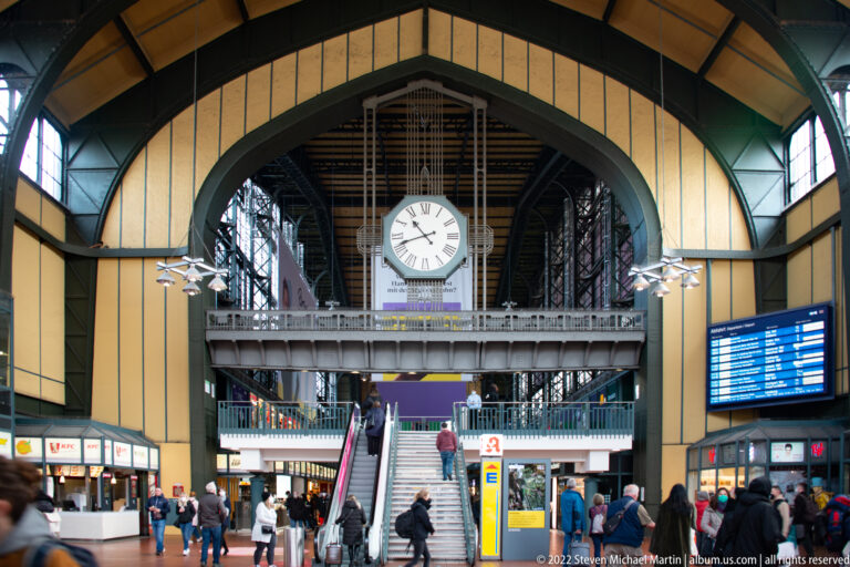 Large round clock above stairs and escalators in the busy interior concourse of Hamburg Hauptbahnhof.