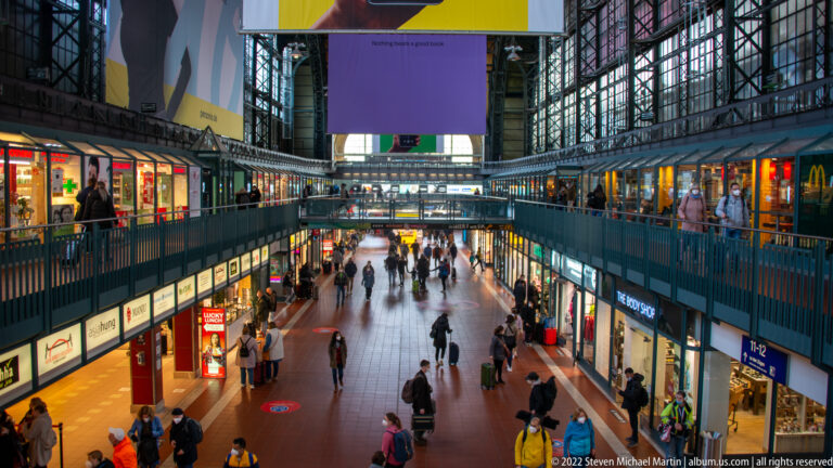 Busy two-level shopping concourse inside Hamburg Hauptbahnhof with shops, travelers, steel roof framing, and suspended signs.