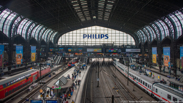 Multiple tracks, platforms, red and white trains beneath the vast arched roof of Hamburg Hauptbahnhof.