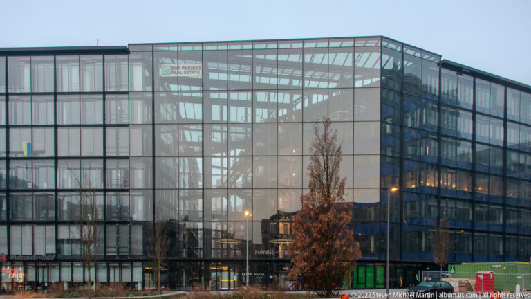 Large modern glass building with visible structural grid, bare tree, and gray overcast sky.