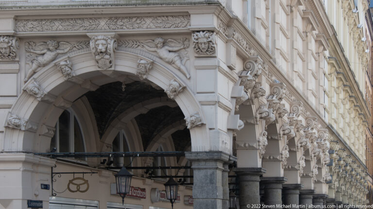 Ornate stone shopping arcade with sculpted masks, repeated carved brackets, hanging lanterns, and storefronts below.