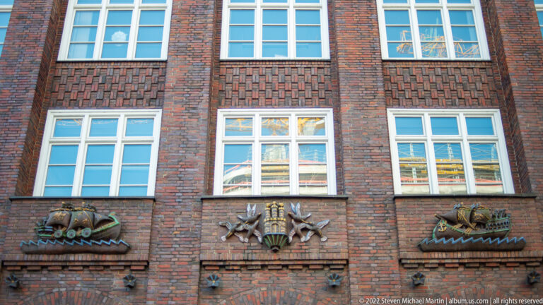 Three colorful maritime and heraldic reliefs mounted across a patterned red-brick façade beneath white windows.