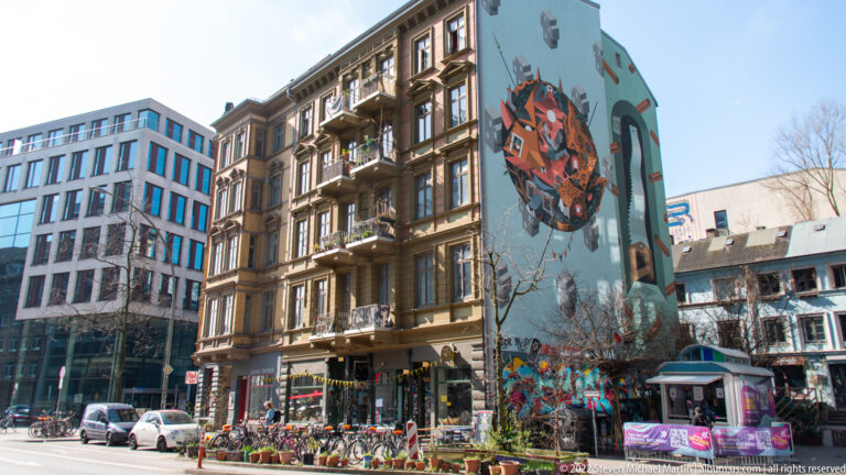 Large colorful mural on the side of an apartment building above street-level graffiti and outdoor displays.