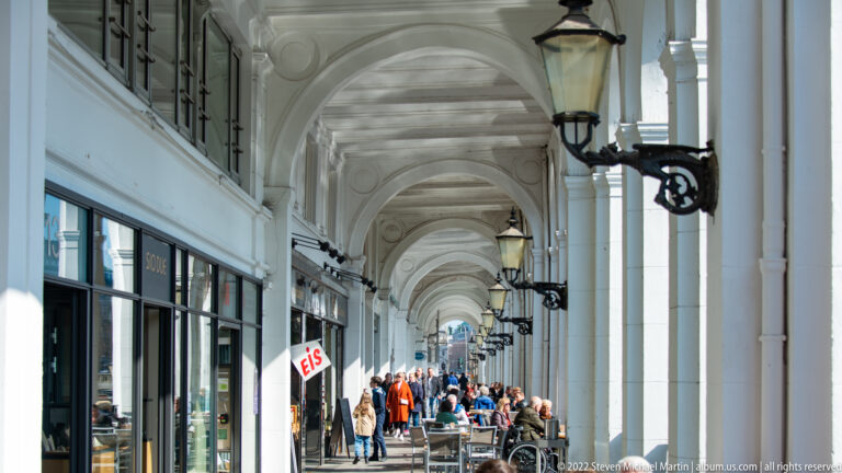 Long white arched shopping colonnade with repeated lamps, storefronts, café seating, and pedestrians.