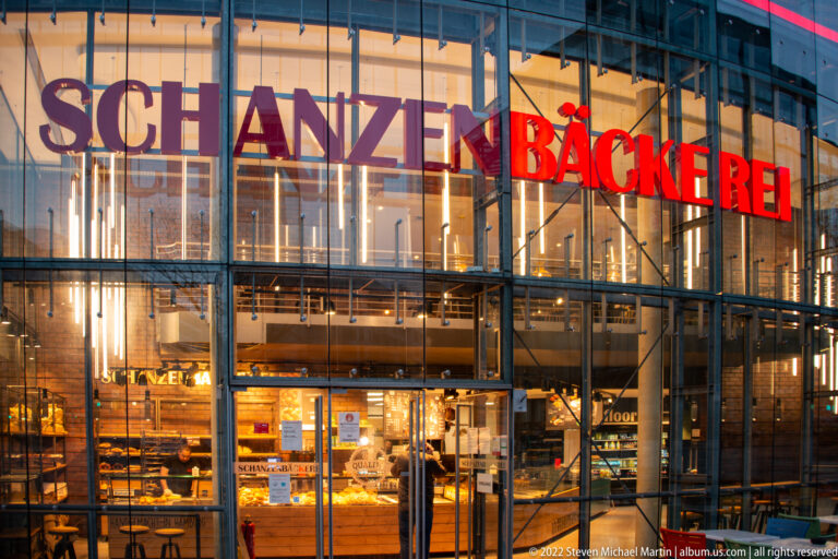 Glass-fronted Schanzenbäckerei with red bakery lettering, illuminated interior, and bread displays behind the windows.