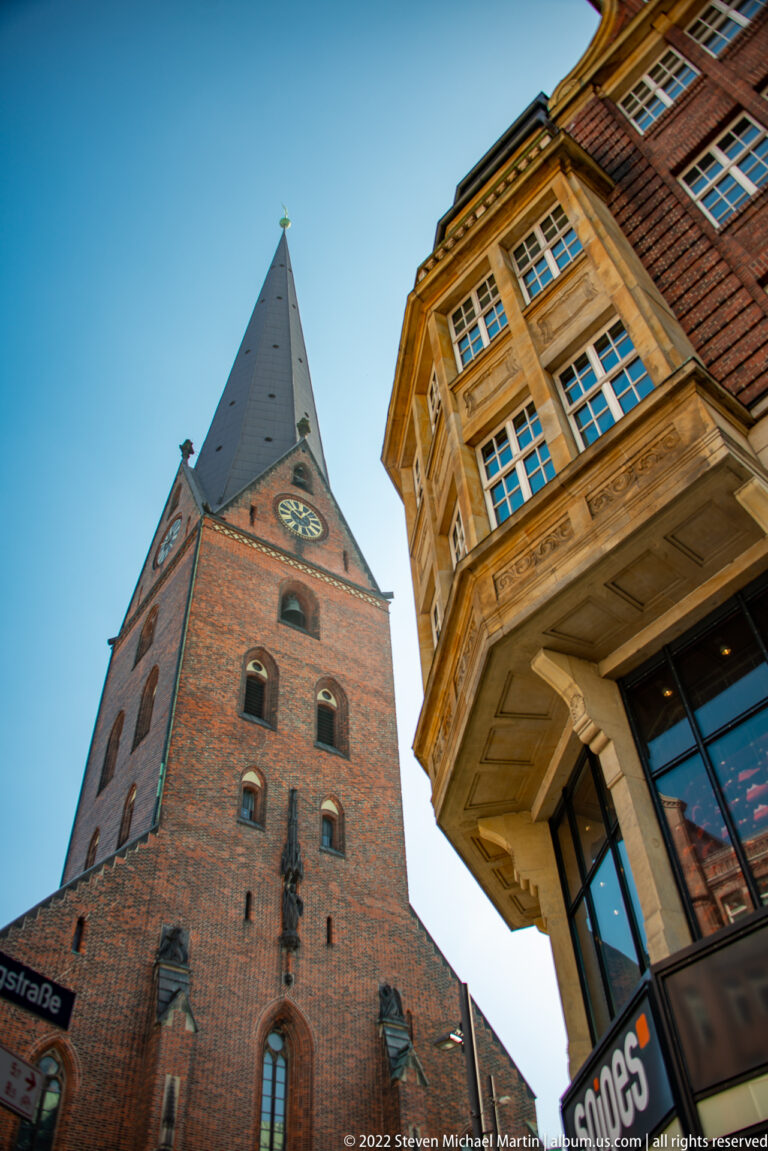 Low-angle view of St. Peter’s Church brick tower with pointed roof and clock faces beside a projecting historic corner building.