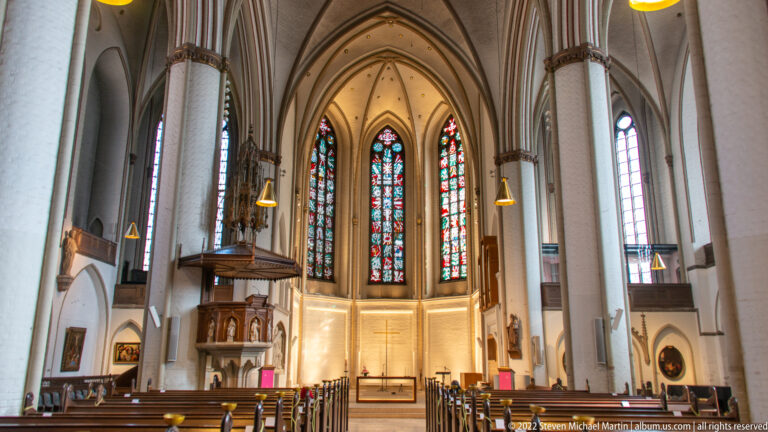 Interior of St. Peter’s Church with tall pale Gothic arches, wooden pews, carved pulpit, and stained-glass windows at the east end.
