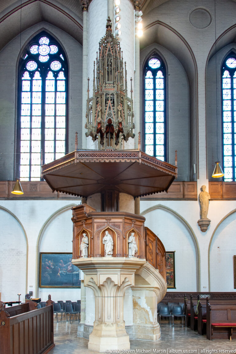 Large carved church pulpit with pale stone base, sculpted wooden panels, Gothic canopy, and tall stained-glass windows behind.