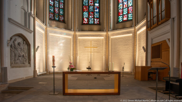 Simple wooden altar in a pale brick chancel with gold cross, tall stained-glass windows, and carved relief at left.
