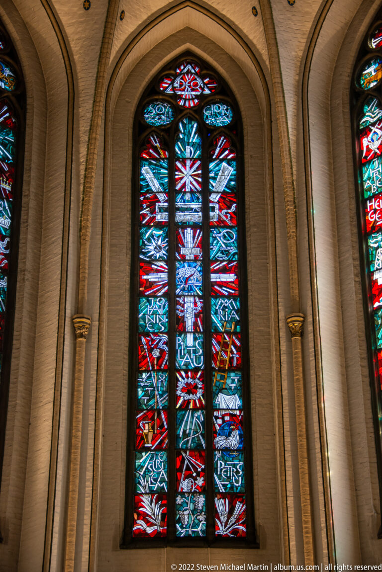 Tall pointed stained-glass window with angular red, blue, white, and black abstract forms in a pale brick Gothic arch.