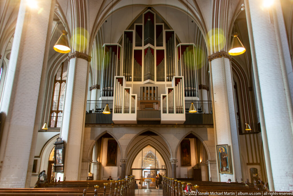 Large pipe organ with stepped silver pipes above pointed arches at the west end of a church nave.