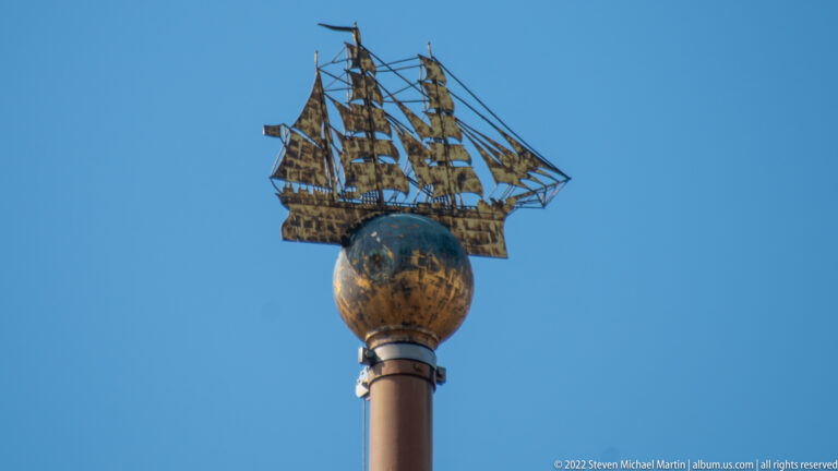 Golden multi-masted sailing ship model mounted above a globe on a tall column against blue sky.