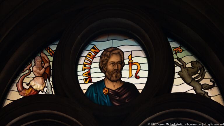 Stained-glass medallion of a bearded male figure labeled “SATURN,” framed by symbolic side panels.