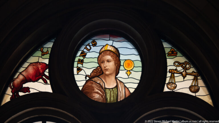 Stained-glass medallion of a crowned female figure labeled “VENUS,” with symbolic side panels.