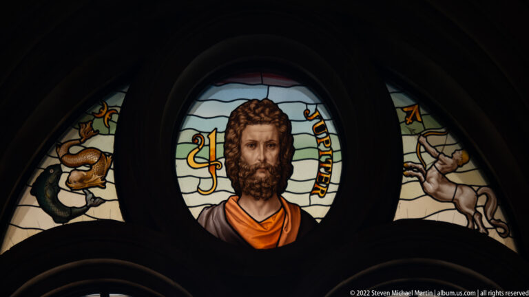 Stained-glass medallion of a bearded male figure labeled “JUPITER,” flanked by symbolic animal and winged figures.