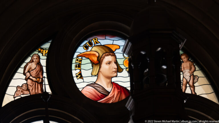 Stained-glass medallion of wing-helmeted Mercury labeled “MERCUR,” with smaller allegorical side figures.