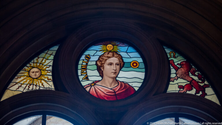 Stained-glass portrait of Apollo labeled “APOLLO,” with radiant sun above and symbolic side panels.