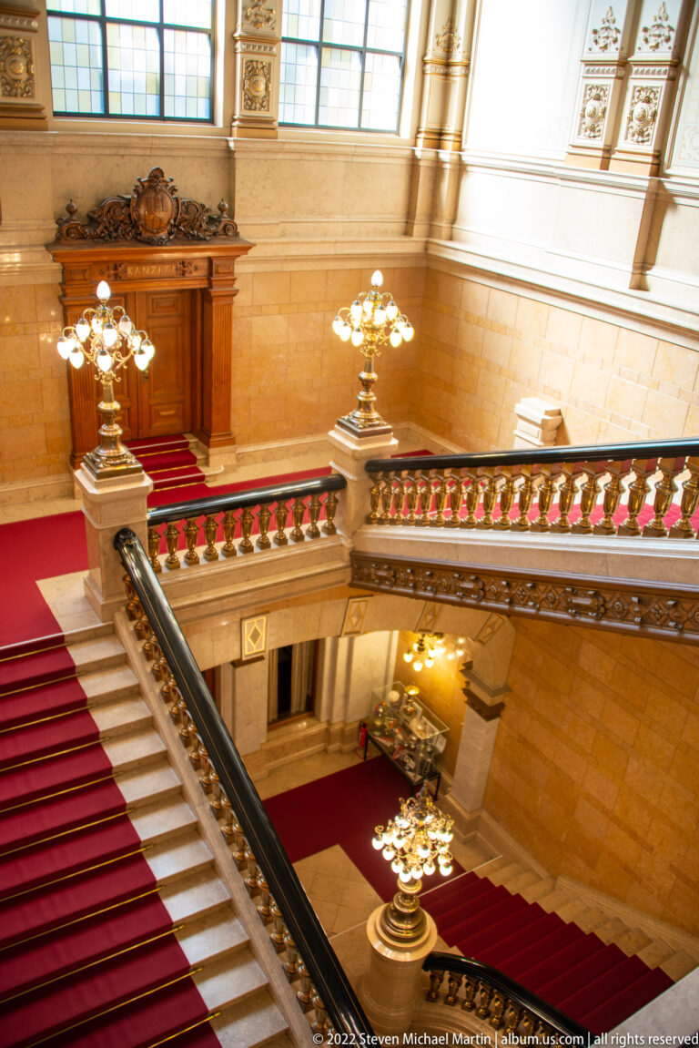 Grand red-carpeted stone staircase inside Hamburg Rathaus with ornate balustrade, arched windows, and brass chandeliers.