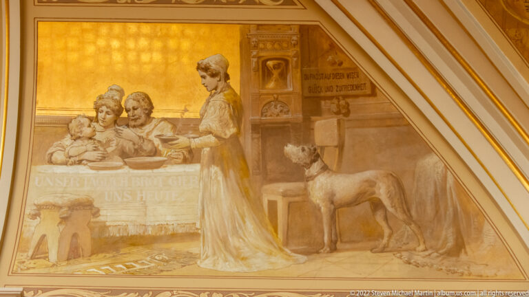Historic Rathaus wall painting with women, children, a dog, and German inscriptions within an ornate gilded frame.