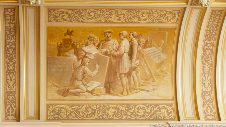 Historic Rathaus mural of men studying large plans amid construction tools and materials, framed by gilded ornament.