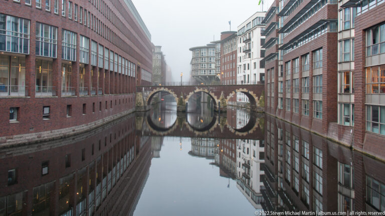 Calm Speicherstadt canal between brick buildings, reflecting a three-arched bridge and fog-softened façades.
