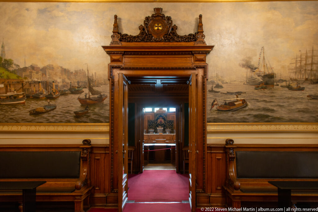 Large harbor mural with boats and sailing ships surrounding an ornate wooden doorway inside Hamburg Rathaus.