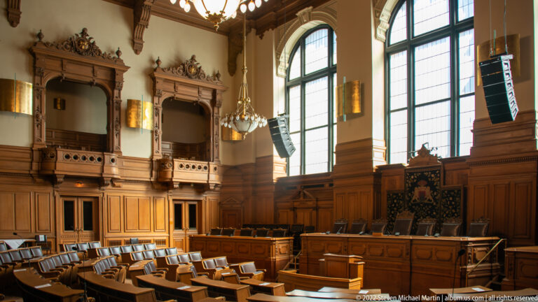 Wood-paneled Hamburg Rathaus chamber with rows of seats, tall arched windows, elevated central chair, and modern speakers.