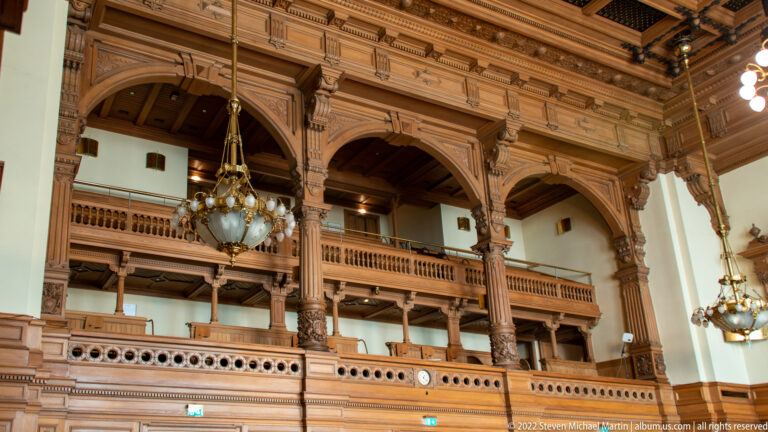 Ornate carved wooden gallery with large supporting columns, balustrades, and chandeliers inside Hamburg Rathaus.