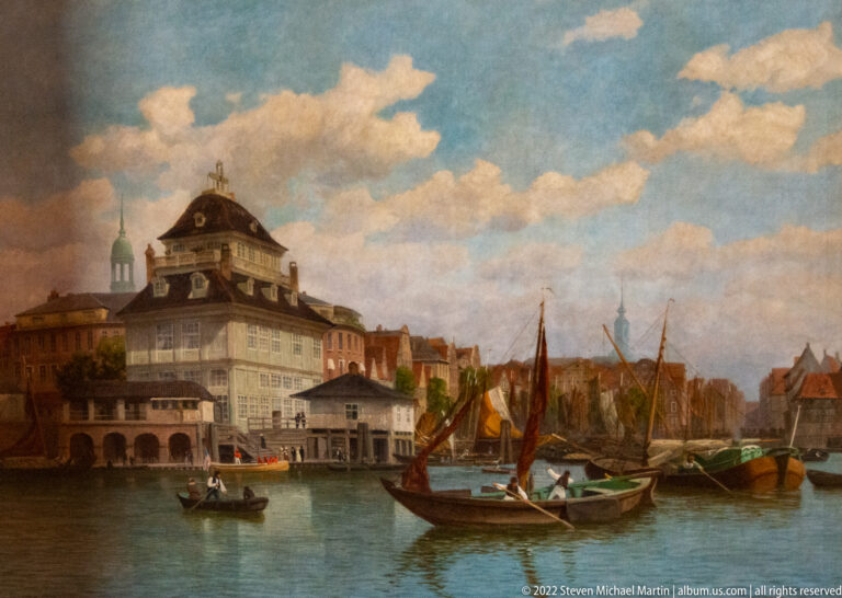 Historic harbor painting with sailing boats, rowboat, waterfront buildings, and broad blue sky.