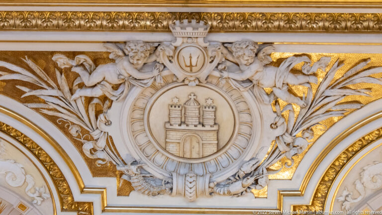 Hamburg castle coat of arms in a white stucco medallion surrounded by gilded foliage, winged figures, and a trident.