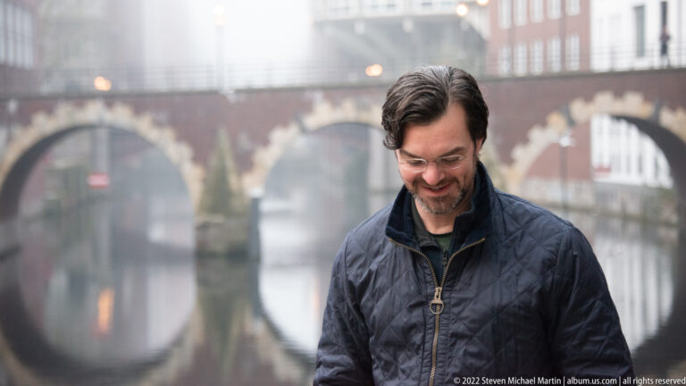Man in dark jacket smiling beside a foggy canal, with a brick arch bridge blurred in the background.