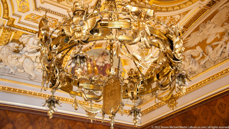 Large gilded sculptural chandelier crowded with figures, floral ornament, shields, and lamps beneath an ornate Rathaus ceiling.