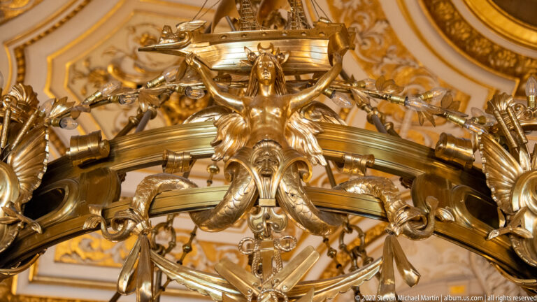 Gilded female figure with raised arms integrated into curling metalwork on an ornate Hamburg Rathaus chandelier.