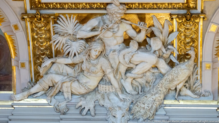 White sculptural group with feathered figures, tropical foliage, reptile, and animal skin against a gilded architectural frame.