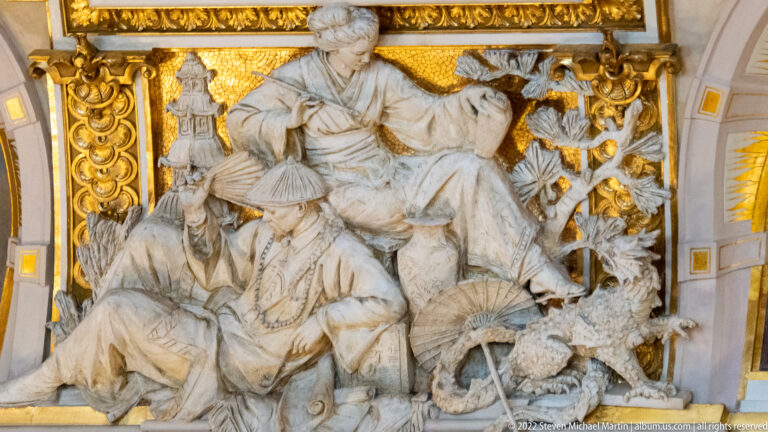 White sculptural group with figures in Asian-style dress, parasol forms, dragon-like creature, and gilded ornament.