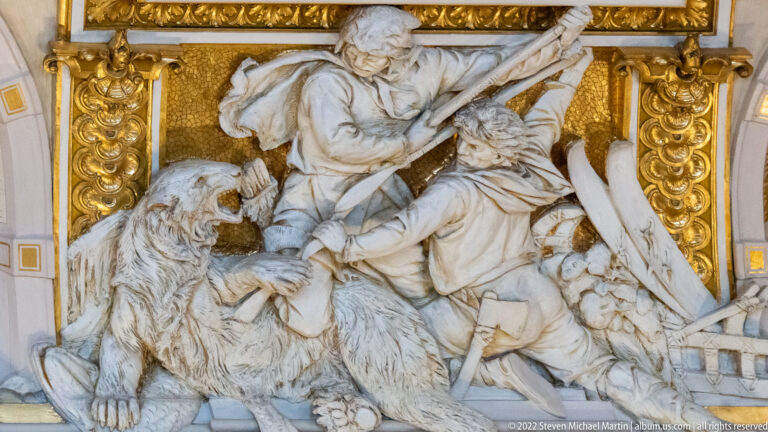White sculptural group with fur-clad figures and a large bear framed by gilded architectural ornament.