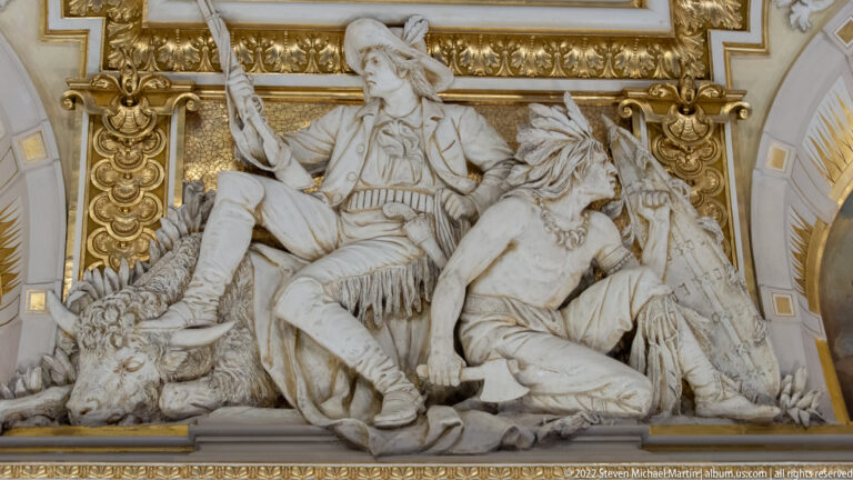 White sculptural relief with seated European-dressed figure, horned animal, and feathered or fringed figure below.