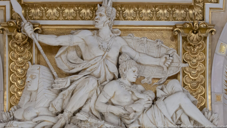 White sculptural relief with Egyptianizing headdress, standing figure, reclining figures, and gilded frame.