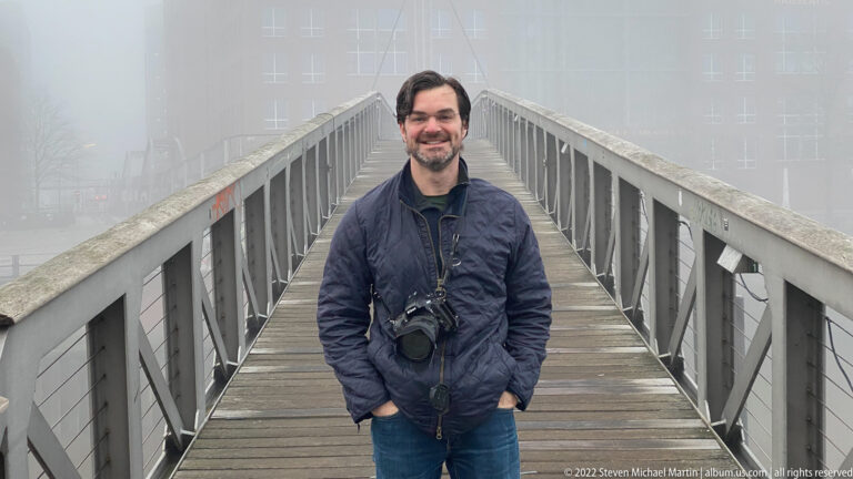 Adult man smiling on a wooden pedestrian bridge as metal railings and modern buildings disappear into dense fog.