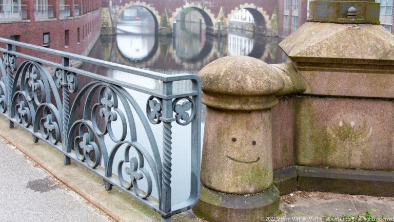 Rounded stone bollard marked with a simple smiley face beside an ornate bridge railing and canal.