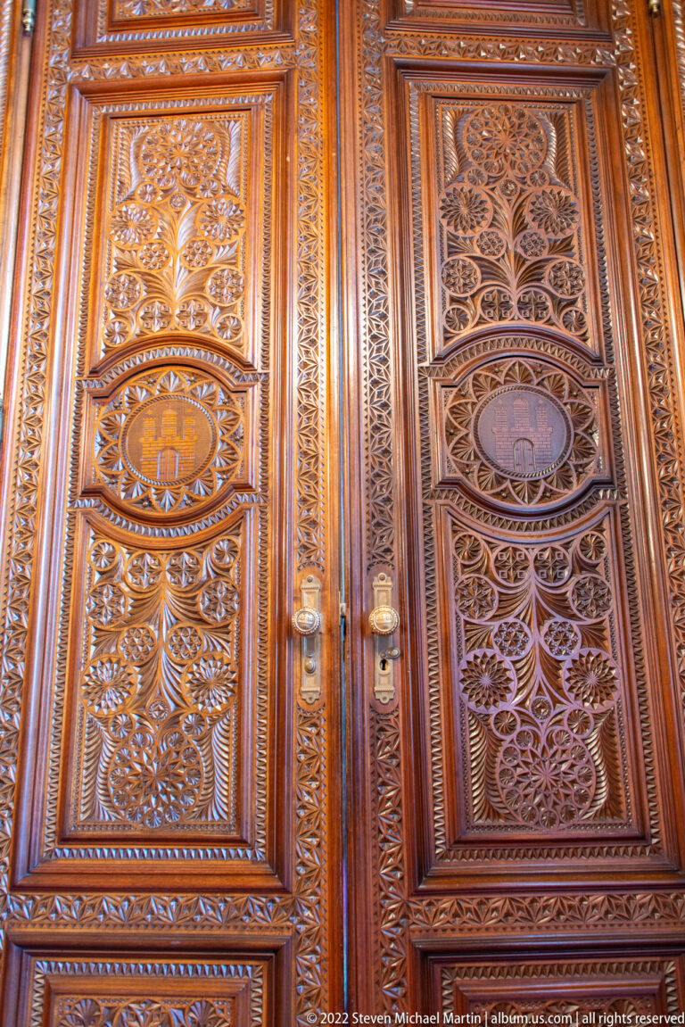 Polished wooden double doors densely carved with foliate openwork, geometric borders, and oval heraldic medallions.