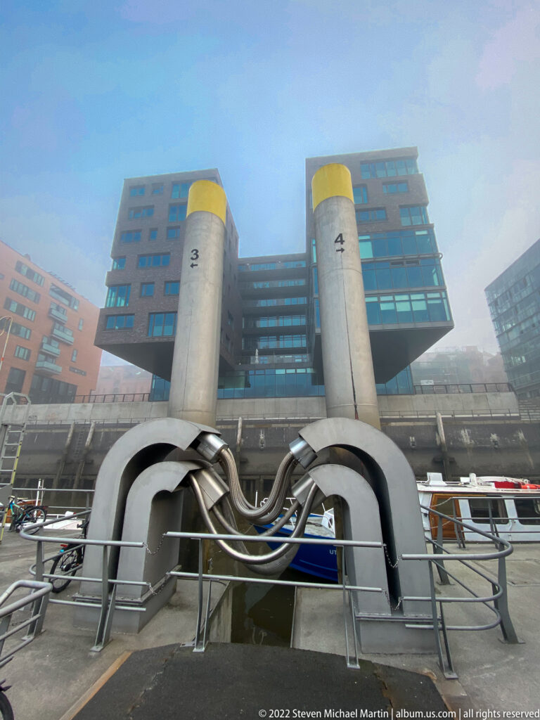 Twin concrete towers numbered 3 and 4 with yellow caps and sweeping dark steel forms beside a modern waterfront building.